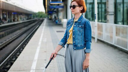 a blind young lady wearing black sunglasses and holding a white cane at a railway station.