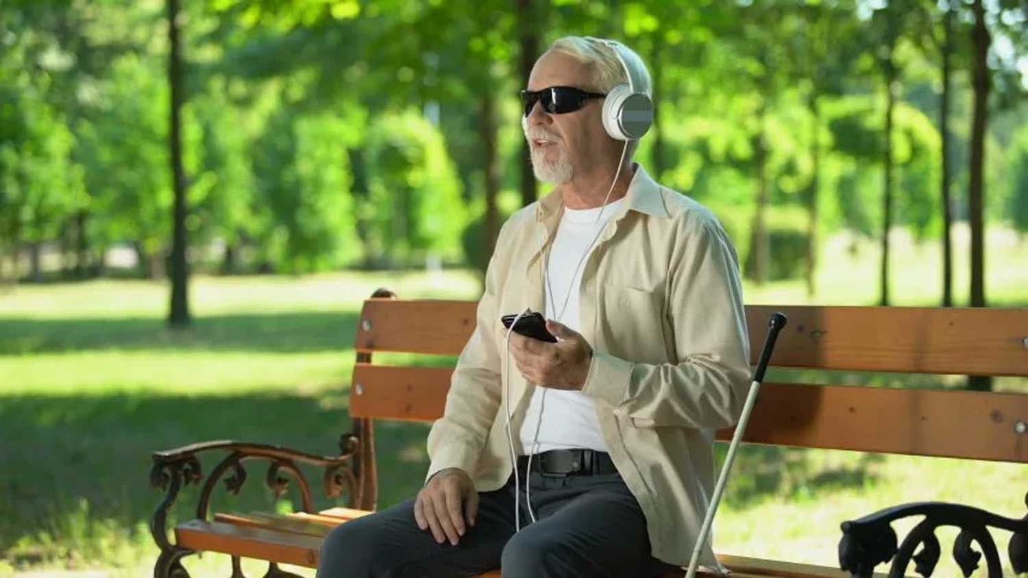 A blind man listening to an audio book on his mobile phone in sitting in a park.