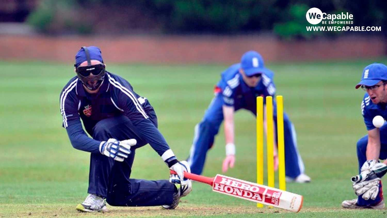 The image shows a game of blind cricket being played. There is a batsman in front of yellow stumps, a wicket-keeper and a fielder.